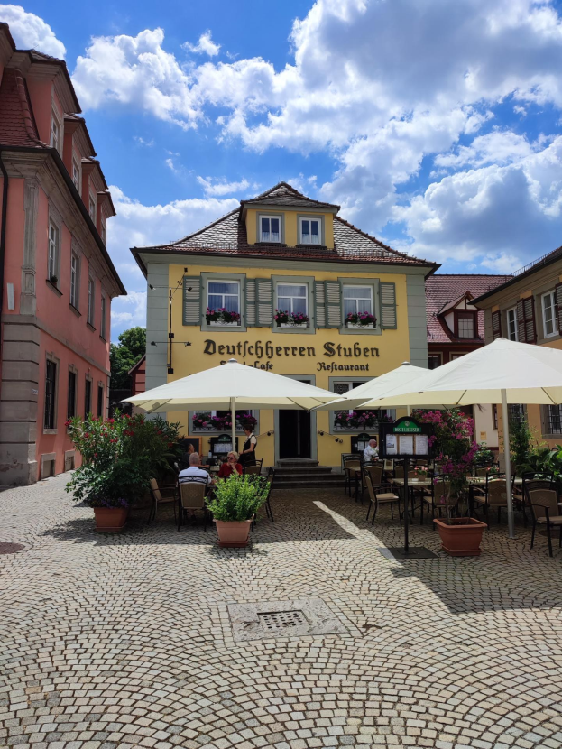 Freisitzbereich mit Tischen und Sonnenschirmen vor einem gelben Gebäude mit der Aufschrift "Deutschen Stuben Restaurant" in Baden-Württemberg, Deutschland, mit einigen Menschen, die sitzen und stehen, unter einem blauen und weißen Himmel.