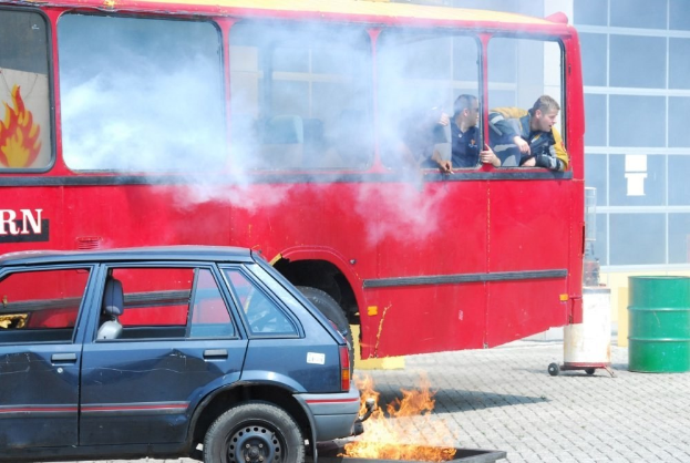 Roter Doppeldeckerbus mit Rauch und einem geparkten Auto daneben, drei sichtbare Passagiere im Inneren und ein Gebäude mit Glasfenstern und einem Fass im Hintergrund.