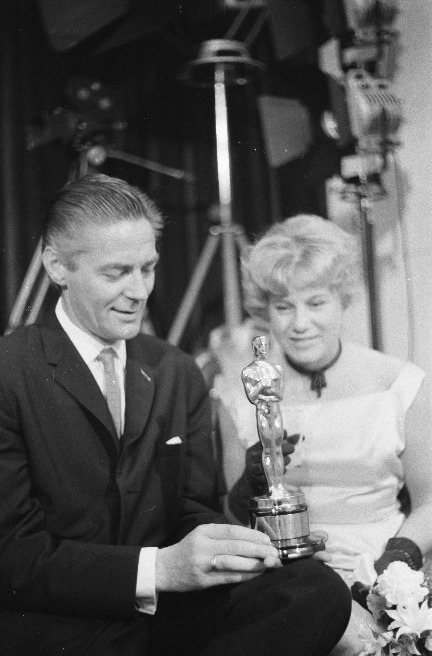 Ein Mann in einem schwarzen Blazer, weißem Hemd und Krawatte hölt eine Oscar-Statue, während er neben einer Frau in Abendgarderobe sitzt, die einen Blumenstraußl in der Hand hält, mit Hintergrundbeleuchtung in einem Schwarz-Weiß-Bild.