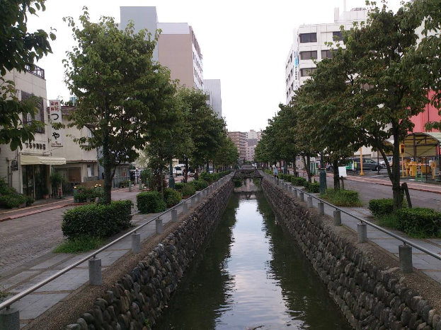 Ein Wasserkanal verläuft durch die Mitte des Bildes, gesäumt von Hecken, Bäumen und einem Fußweg auf beiden Seiten, mit Straßen, Gebäuden und fahrenden Autos im Hintergrund unter einem Himmel.