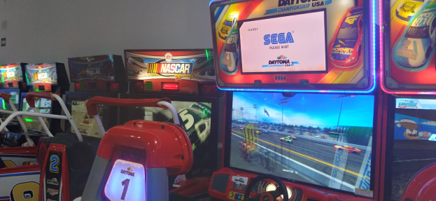 Eine Reihe buntfarbiger Arcade-Automaten, hauptsächlich Rennwagen-Spiele, in einem Raum mit einer Wand im Hintergrund.