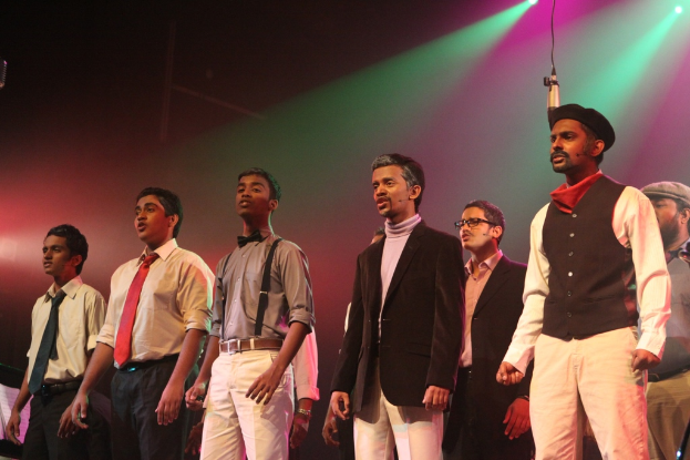 Eine Gruppe von Männern auf einer Bühne, die ein Drama aufführen, mit grünen Scheinwerfern im Hintergrund.