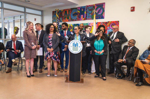 Gruppe von Menschen vor einem Rednerpult mit Mikrofon und einer Tafel mit Text, einige sitzen auf Stühlen und andere stehen, mit Fahnen, Feuerlöscher und Deckenleuchten an der Rückwand.