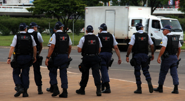 Eine Gruppe von Beamten in Uniform mit M√ľtzen, Hemden, Schutzwesten, Uhren, Hosen und Schuhen, die auf einer Stra√e mit einem geparkten Lkw rechts neben einem Zaun, Geb√§uden, B√§umen, Pflanzen, Masten und Bannern im Hintergrund und einer Br√ľcke oben rechts gehen.