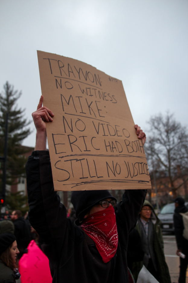 Eine Gruppe von Menschen, die auf einer Straße protestieren, wobei ein Mann ein Schild mit Text hält, Bäume, ein Auto und einen Himmel im Hintergrund.