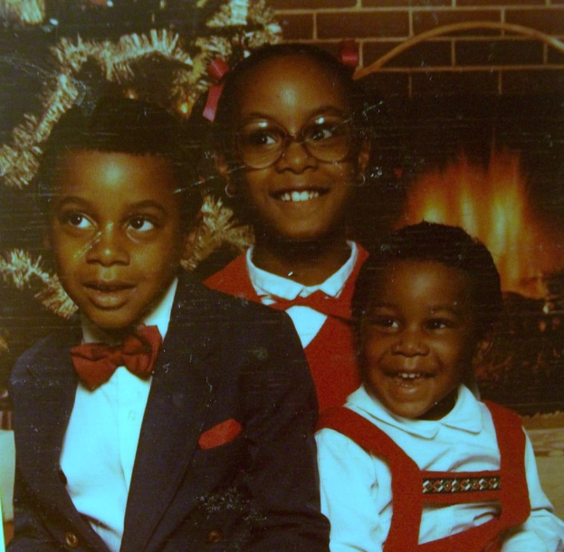 Drei Kinder, die vor einem Weihnachtsbaum lachen, mit einem Kamin aus Holz auf der rechten Seite.