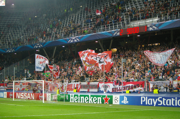 Ein Fußballspiel wird in einem gut beleuchteten Stadion mit einer großen Zuschauermenge, grünem Rasen, einem zentralen Torpfosten mit Bannern und einem Bildschirm in der oberen linken Ecke gespielt.