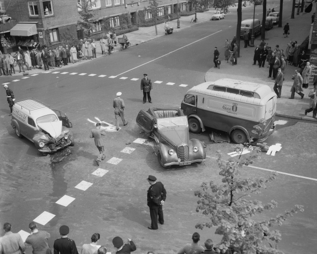 Schwarz-weiß-Szene eines Autounfalls an einer Straßenkreuzung mit einem Van, anderen Fahrzeugen und einer Gruppe von Menschen in der Nähe, mit Gebäuden, Bäumen, Polen und Texttafeln im Hintergrund.