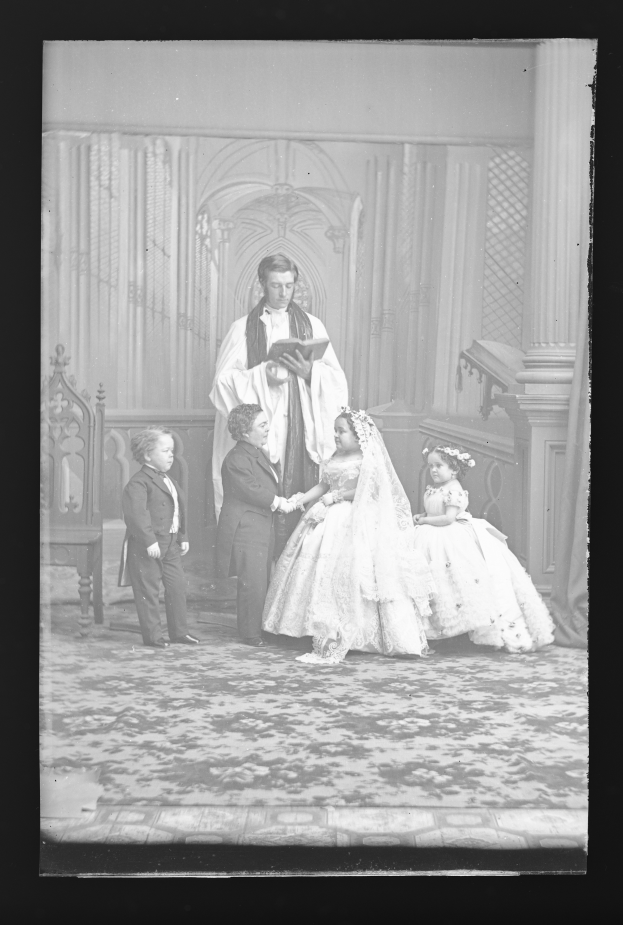 Ein altes Schwarz-Weiß-Foto einer Braut in einem weißen Kleid und Schleier, eines Bräutigams mit einem Buch in der Hand und ihren Kindern vor ihnen, mit einer Wand im Hintergrund.