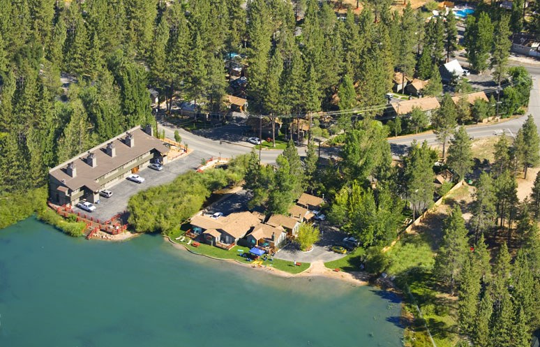 Luftaufnahme eines Resorts am Seeufer mit umliegenden Bäumen, Häusern, Fahrzeugen, Straßen, Masten und Gras, mit dem See unten sichtbar.