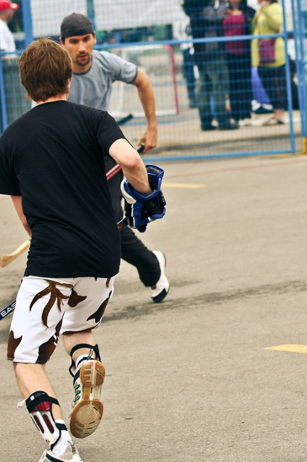 Zwei Personen spielen Hockey auf einer Straße mit einem Zaun und einer Zuschauermenge im Hintergrund.