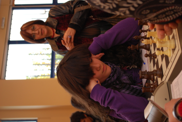 Ein Mädchen in einem lila T-Shirt sitzt beim Schachspielen, während eine Frau in einer Jacke zusieht, mit einer Glastür und einer gelben Wand im Hintergrund.