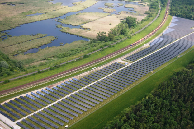 Luftaufnahme einer großen Solar-Farm in einem Feld mit Bäumen, Gras, Pflanzen, Wasser, Solarpanelen auf dem Boden und einem Zug, der entlang einer Bahnstrecke fährt.
