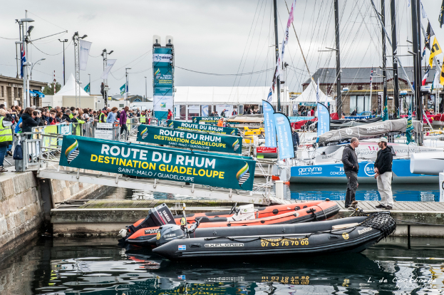 Eine belebte Wasserfrontszene mit zahlreichen Booten, Bannern, Pfählen und Drähten, einer großen Menge hinter einer Absperrung auf der linken Seite und zwei Personen, die auf einer Brücke über dem Wasser stehen, mit Abteilen um die Boote herum.