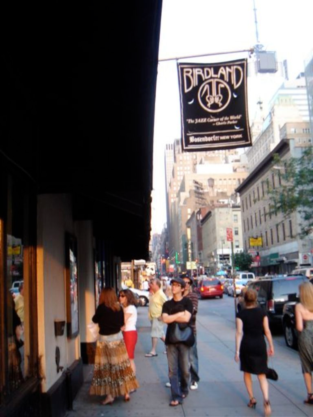 Eine belebte Straßenszene mit zahlreichen Fahrzeugen, Gebäuden auf beiden Seiten, Fußgängern, die die Straße entlanggehen, und einer Gruppe von Menschen, die vor einem Laden auf der linken Seite stehen.