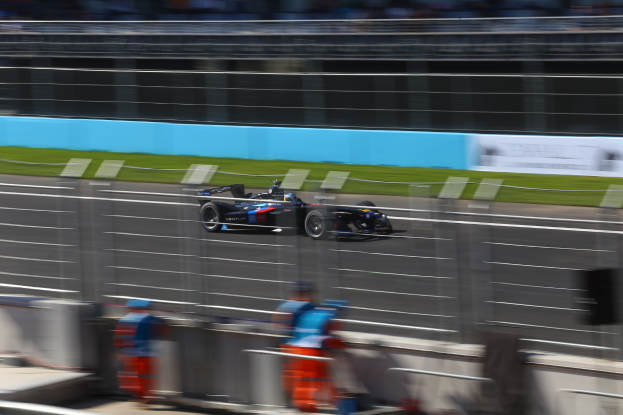 Ein Formel-Auto fährt auf einer Rennstrecke an einer Menge von Zuschauern vorbei, Gras auf dem Boden und Schilder im Hintergrund.