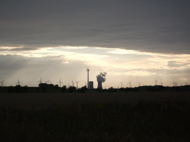 Kraftwerk mit Windrädern vor Wolkenhimmel, umgeben von Grün am Boden.