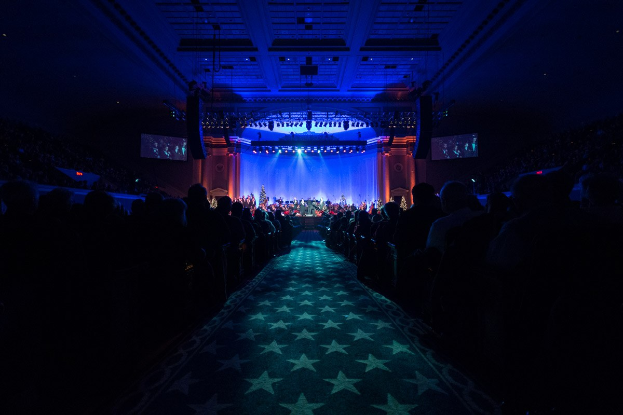 Ein großer Auditorium mit sitzenden Zuschauern, eine Bühne mit Musikern und überkopfbeleuchtung und -bildschirme, was auf einen Konzertsaal während der Nationalen Convention 2019 hindeutet.