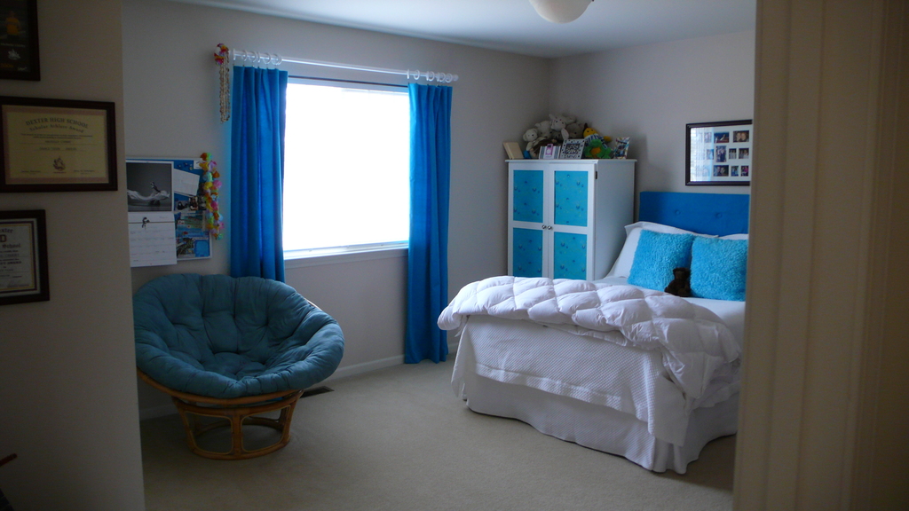 Ein Raum mit einem Bett, einem Stuhl, einem Fenster mit Vorhängen, einem Schrank, einem Foto Rahmen und einem gefliesten Boden.