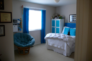 Ein Raum mit einem Bett, einem Stuhl, einem Fenster mit Vorhängen, einem Schrank, einem Foto Rahmen und einem gefliesten Boden.