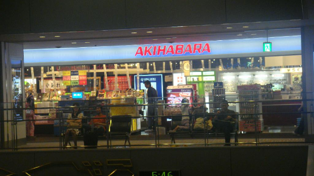 Ein Geschäft mit zwei Bänken und einigen Menschen davor, eine Wand mit Text darüber, verschiedene Gegenstände in Schalen drinnen und eine eingezäunte Wand unten.