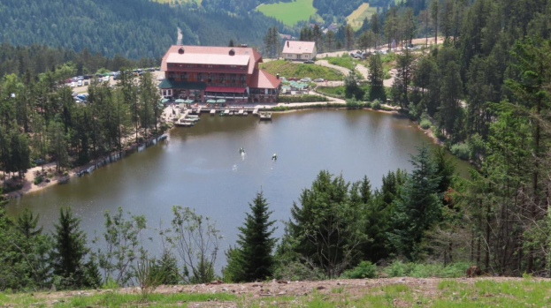 Großer See umgeben von Bäumen und Bergen, mit Gebäuden, Fahrzeugen und Menschen im Vordergrund, Hügel und einen klaren blauen Himmel im Hintergrund, saftiges Grün im Wasser und Pflanzen am Boden und eine Lodge in der Mitte des Sees.