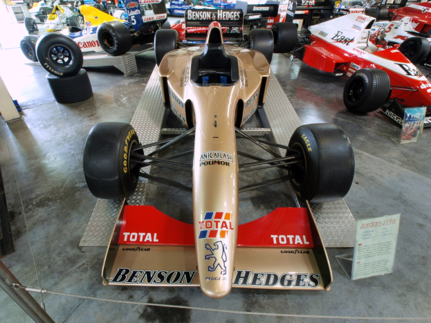 Eine Gruppe von Rennwagen in einem Motorsportmuseum ausgestellt, getrennt durch einen Metallzaun auf der linken Seite, mit lesbarem Informationstext im Hintergrund.