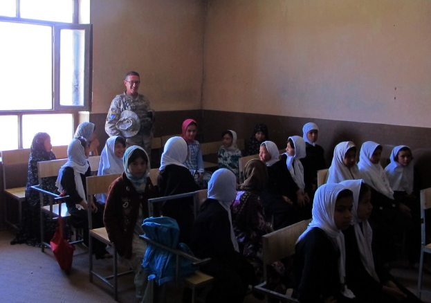 Eine Klasse mit Schülern auf Bänken im Vordergrund, ein strahlender Militärmann dahinter und eine braune Wand mit Glasfenster im Hintergrund.