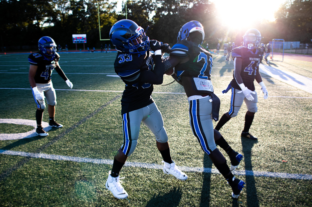 Gruppe junger Männer spielt Fußball auf einem Feld mit Bäumen und einem klaren blauen Himmel, trägt Helme und Schuhe.