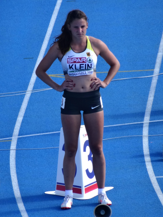 Eine Frau steht auf einer blauen Bahn und hat ihre Hände in den Hüften, trägt eine Sportuniform mit Shorts und Schuhen.