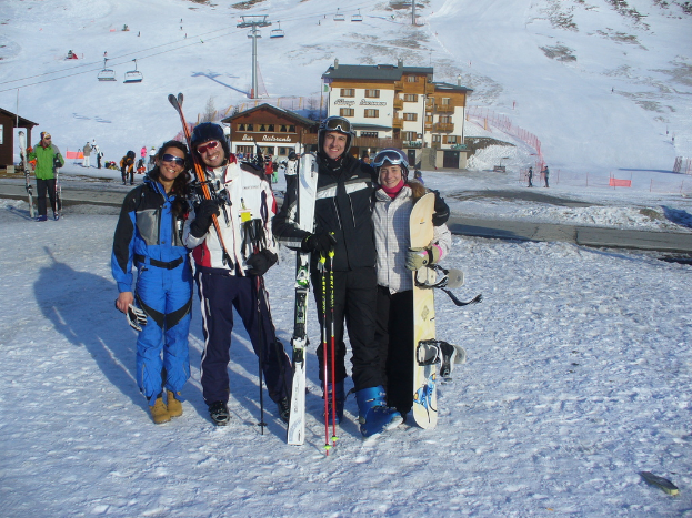 Vier Personen posieren für ein Foto mit Skaterpads und Werkzeugen, mit Gebäuden und anderen Menschen im Hintergrund, vor einer schneebedeckten und vereisten Berglandschaft.