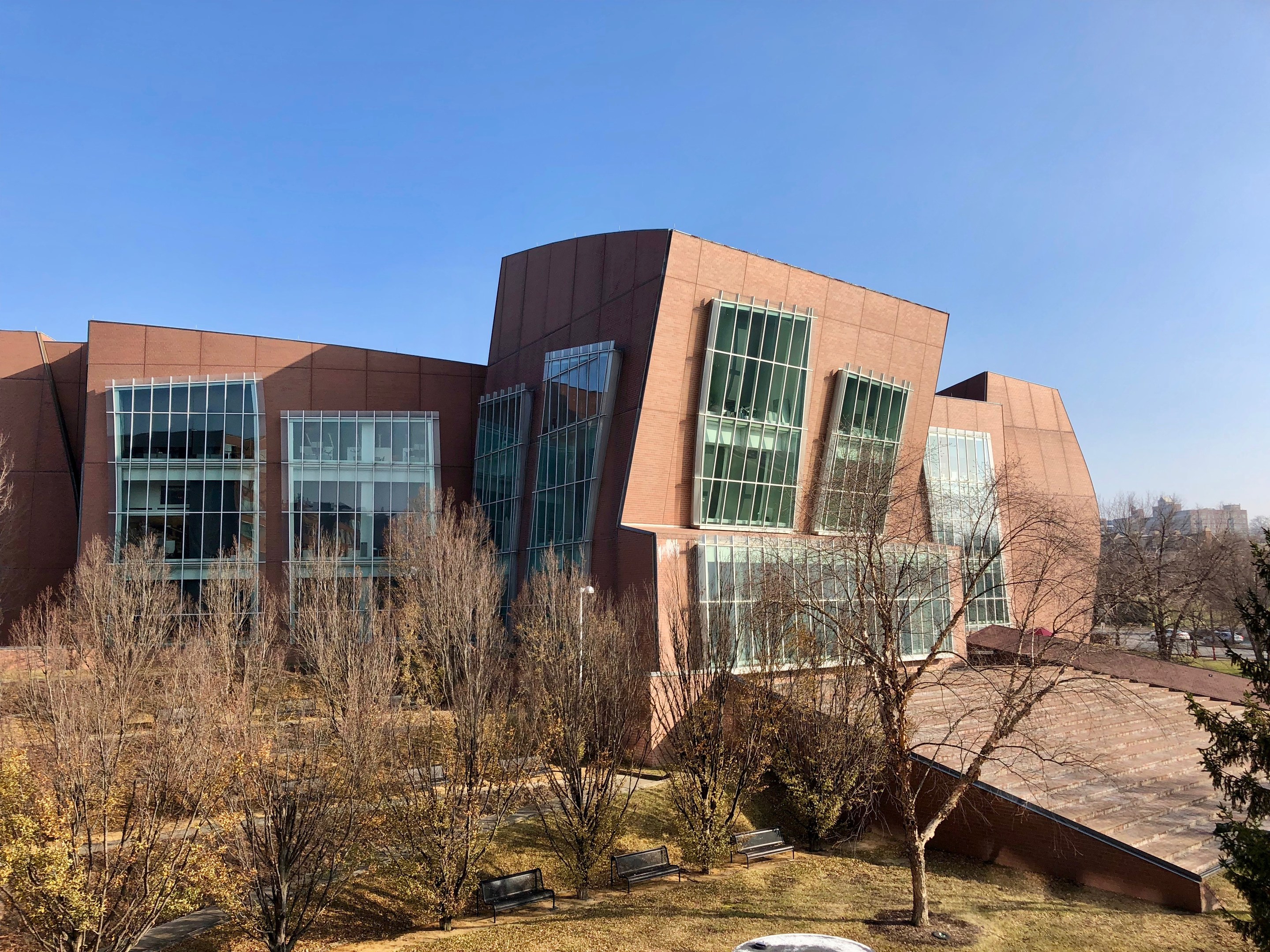 Großes Gebäude mit vielen Fenstern, umgeben von Bäumen, Bänken, Gras und einem Laternenpfahl, mit anderen Gebäuden und einem klaren blauen Himmel im Hintergrund; als der neue Campus der University of Illinois at Chicago bezeichnet.