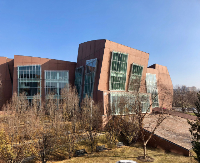 Großes Gebäude mit vielen Fenstern, umgeben von Bäumen, Bänken, Gras und einem Laternenpfahl, mit anderen Gebäuden und einem klaren blauen Himmel im Hintergrund; als der neue Campus der University of Illinois at Chicago bezeichnet.