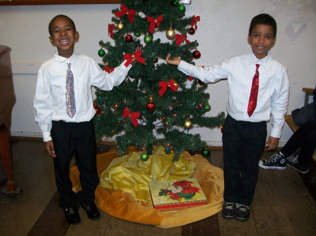 Ein geschmückter Weihnachtsbaum steht in einem Raum mit einer Kiste, zwei Gegenständen in der Nähe, einem Holztisch links, einem Stuhl rechts, einer weißen Wand mit angebrachten Gegenständen im Hintergrund und zwei Jungen in weißen Hemden, die lächeln und den Baum halten.