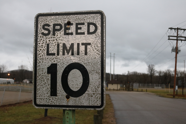 Ein Tempolimit-Schild am Straßenrand, umgeben von Gras, Strommasten mit Kabeln, einem Zaun, Häusern, Bäumen und einem bewölkten Himmel im Hintergrund.