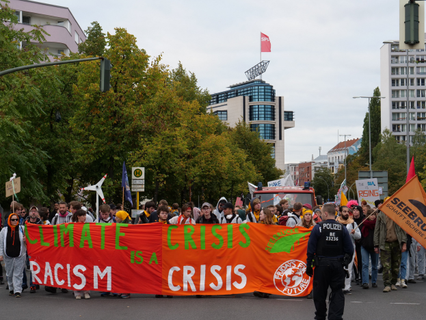 Eine Gruppe von Menschen marschiert eine baumbestandene Straße entlang und hält ein Banner mit der Aufschrift "Klima-Krise ist eine Krise", mit Gebäuden und einem klaren blauen Himmel im Hintergrund.