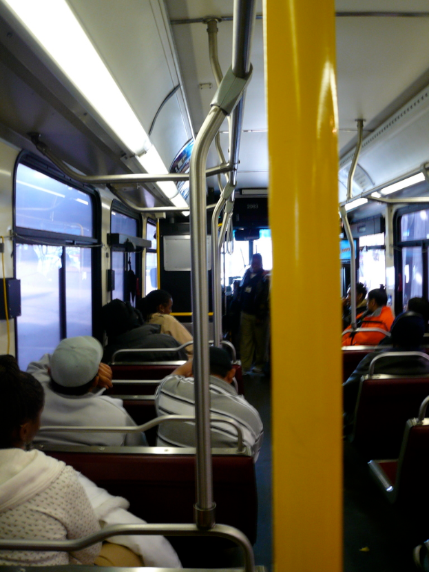 Innenansicht eines Busses: Passagiere sitzen auf Bänken mit vertikalen Stangen neben einigen Sitzen.