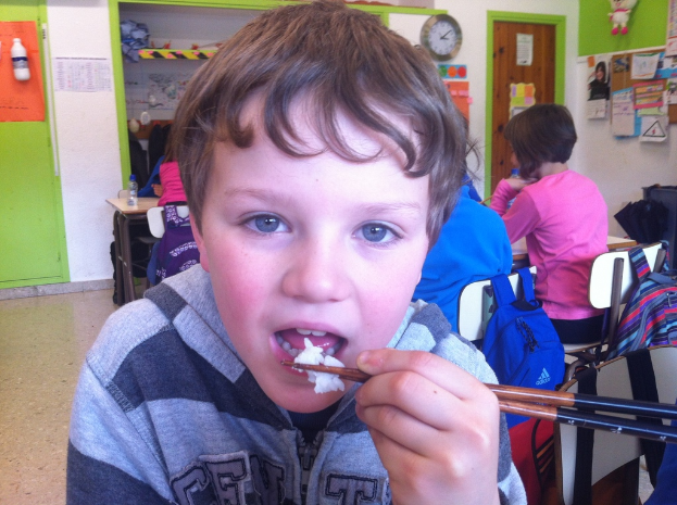 Ein Junge in einer grauen und schwarzen Jacke isst mit Stäbchen in einem Klassenzimmer, mit anderen Kindern hinter ihm, einer grün-weißen Wand, Lehrplakaten und Diagrammen an einer weißen Wand, einer braunen Tür und einer Wanduhr.