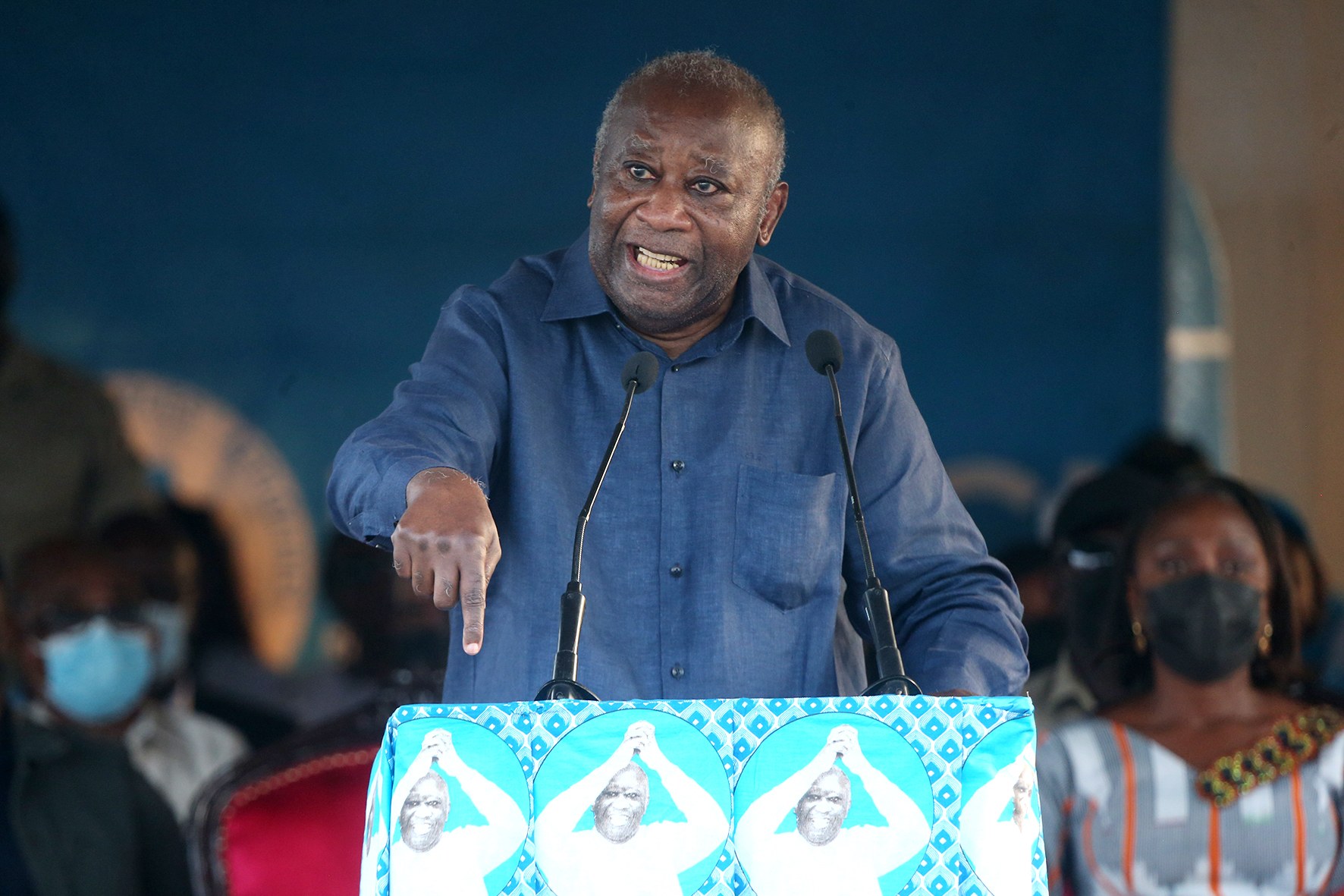 Ein Mann in einem blauen Hemd spricht an einem Pult mit zwei Mikrofonen, gewandt einem sitzenden Publikum, das Masken trägt, mit einer Wand im Hintergrund.