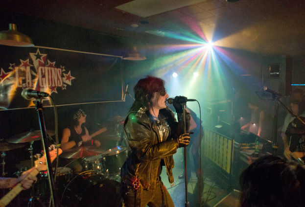 Eine Frau in einer schwarzen Jacke singt auf der Bühne in ein Mikrofon, während ein Mann mit Sonnenbrille und Stirnbinde hinter ihr eine Bassgitarre spielt und ein 'LA Guns'-Plakat sowie Diskoleuchten im Hintergrund zu sehen sind.