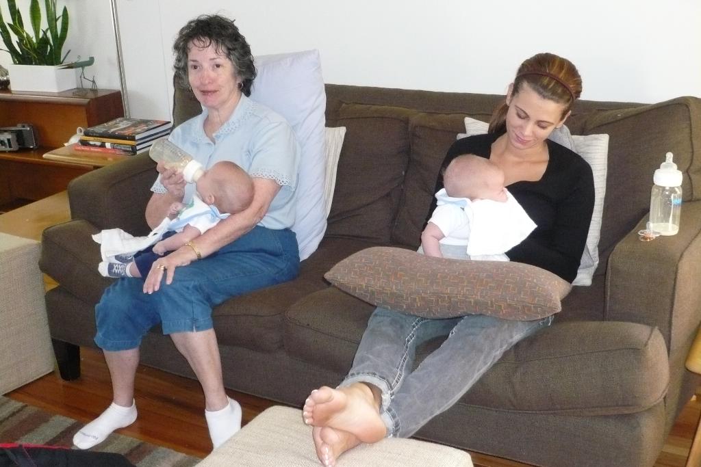 Zwei Frauen sitzen auf einem Sofa und halten Babys, mit Kissen drumherum, vor einer weißen Wand, mit einem Schreibtisch, Büchern und einer Zimmerpflanze links daneben.