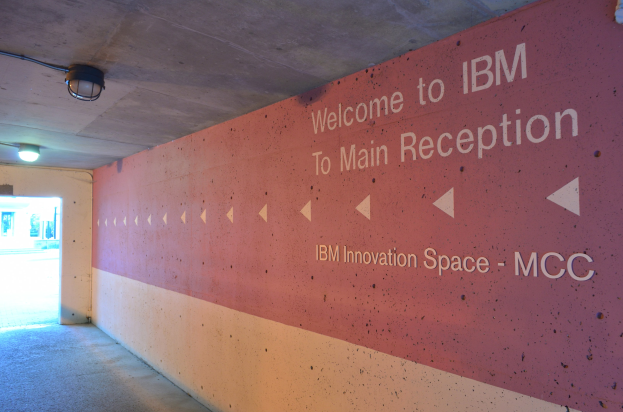 Eine Wand mit den Worten "Willkommen bei IBM" beleuchtet von Deckenlampen, mit einer Tür im Hintergrund beschriftet "IBM Innovation Space MCC".