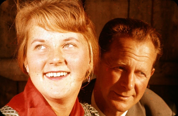 Ein Mann und eine Frau in inniger Umarmung, wobei der Mann seinen Arm um die Taille der Frau gelegt hat und ihr Kopf auf seiner Schulter ruht, vor einer schmucklosen Wand stehend.