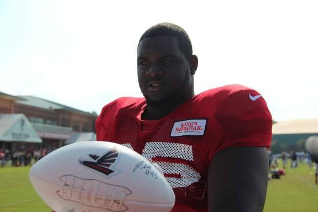 Ein Mann in einem roten Trikot hält einen Ball in der Mitte eines Stadions, mit Gras, einem Gebäude, dem Himmel und einer Menge im Hintergrund.