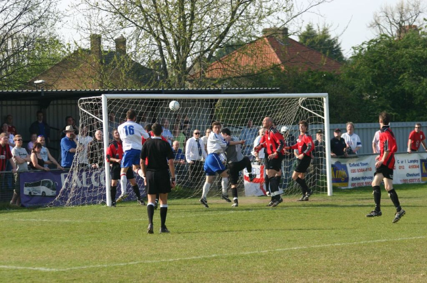 Fußballspieler spielen auf einem Feld mit einem Tor hinter sich, während Zuschauer von der Seitenlinie zuschauen, mit Bäumen und Häusern im Hintergrund.