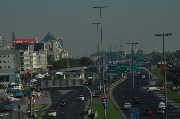 Eine Luftaufnahme einer Stadt mit Gebäuden, Türmen, Kraftfahrzeugen, Werbetafeln, Schildern, Straßen, Bäumen, Straßenlaternen, Menschen, Verkehrskegeln und Informationsanzeigen unter einem klaren Himmel.
