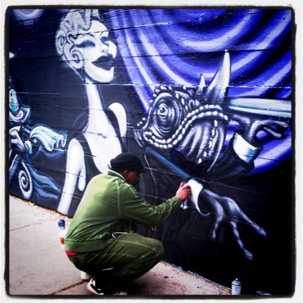 Eine Person hält eine Flasche in der Hand und steht auf einer Straße, umgeben von mehreren anderen Flaschen, mit einer graffiti-bedeckten Wand im Hintergrund.