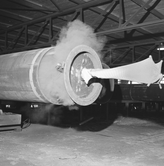 Ein Schwarz-Weiß-Foto einer großen Rohrleitung in einer Fabrik, aus der Rauch aufsteigt, mit einer Maschine links und anderen Gegenständen rechts, unter einem sichtbaren Fabrioddach.