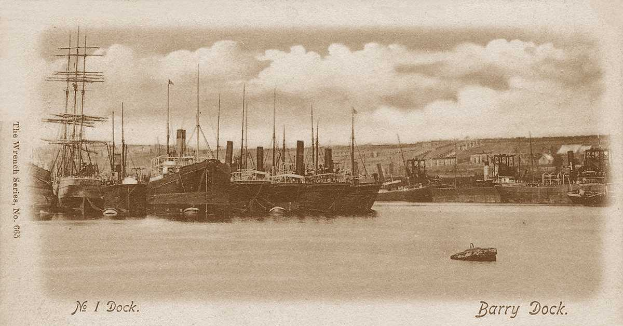 Ein altes Schwarz-Weiß-Foto von Booten, die am Barry Dock in Wrexham, England, liegen, mit Gebäuden im Hintergrund und Wolken am Himmel.