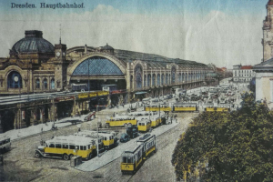 Eine alte Postkarte, die einen belebten Dresdner Bahnhof mit Fahrzeugen, Menschen, Bäumen, Gebäuden und einem bewölkten Himmel zeigt, mit der Aufschrift «Dresden Hauptbahnhof» oben.